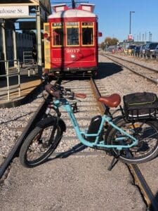 Here's an alt tag for the image: Light blue ebike by train tracks.