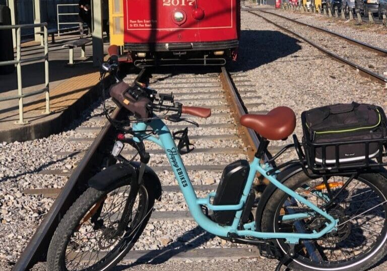 Here's an alt tag for the image: Light blue ebike by train tracks.